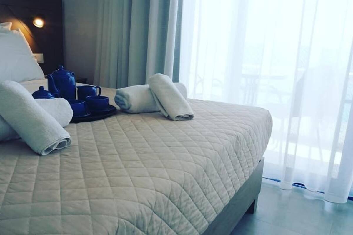 A neatly made bed with rolled towels, a set of blue mugs, and a tea set on the bed.