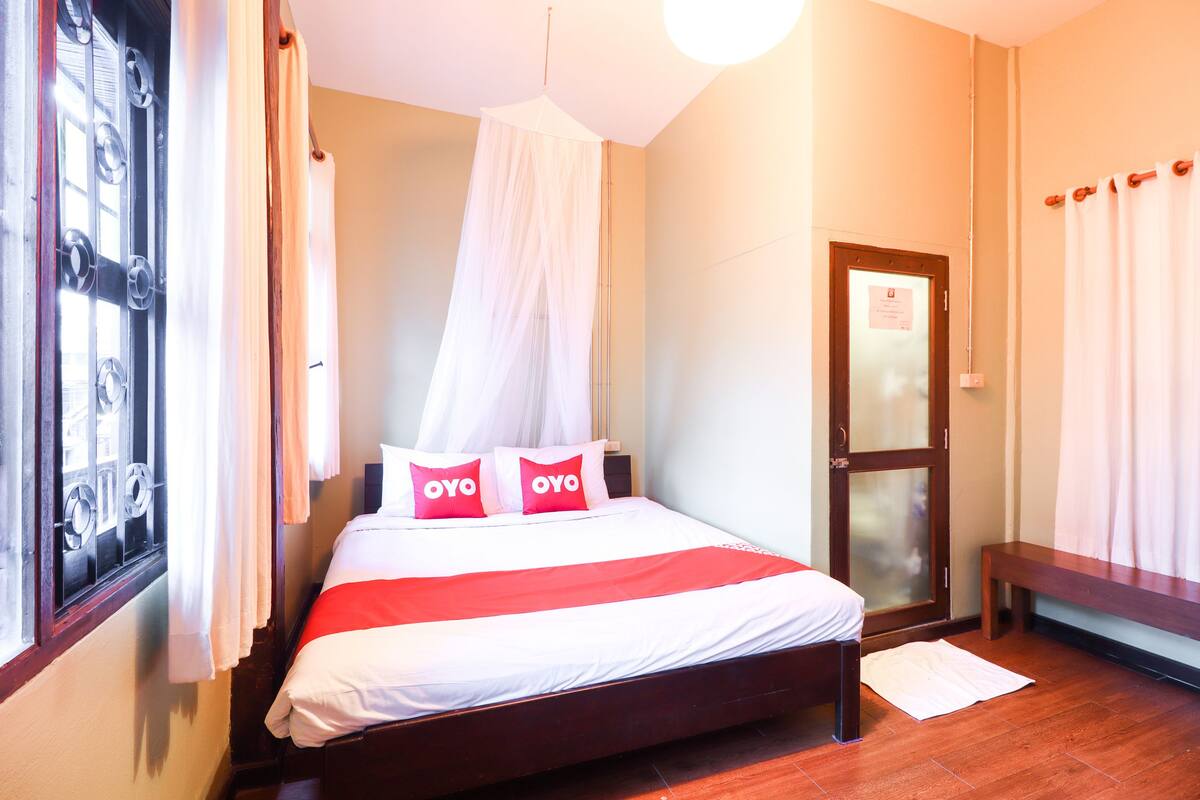 Een net opgemaakt bed met een wit en rood gestreept dekbedovertrek, twee kussens met 'OYO'-branding, een houten hoofdbord en een houten bankje naast het bed.