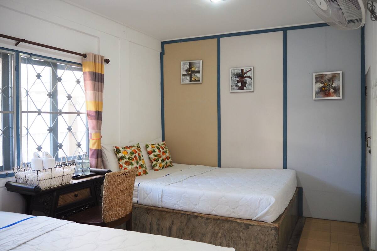 A bedroom with a bed, a window with a curtain, a wall with three framed pictures, and a small table with a basket.