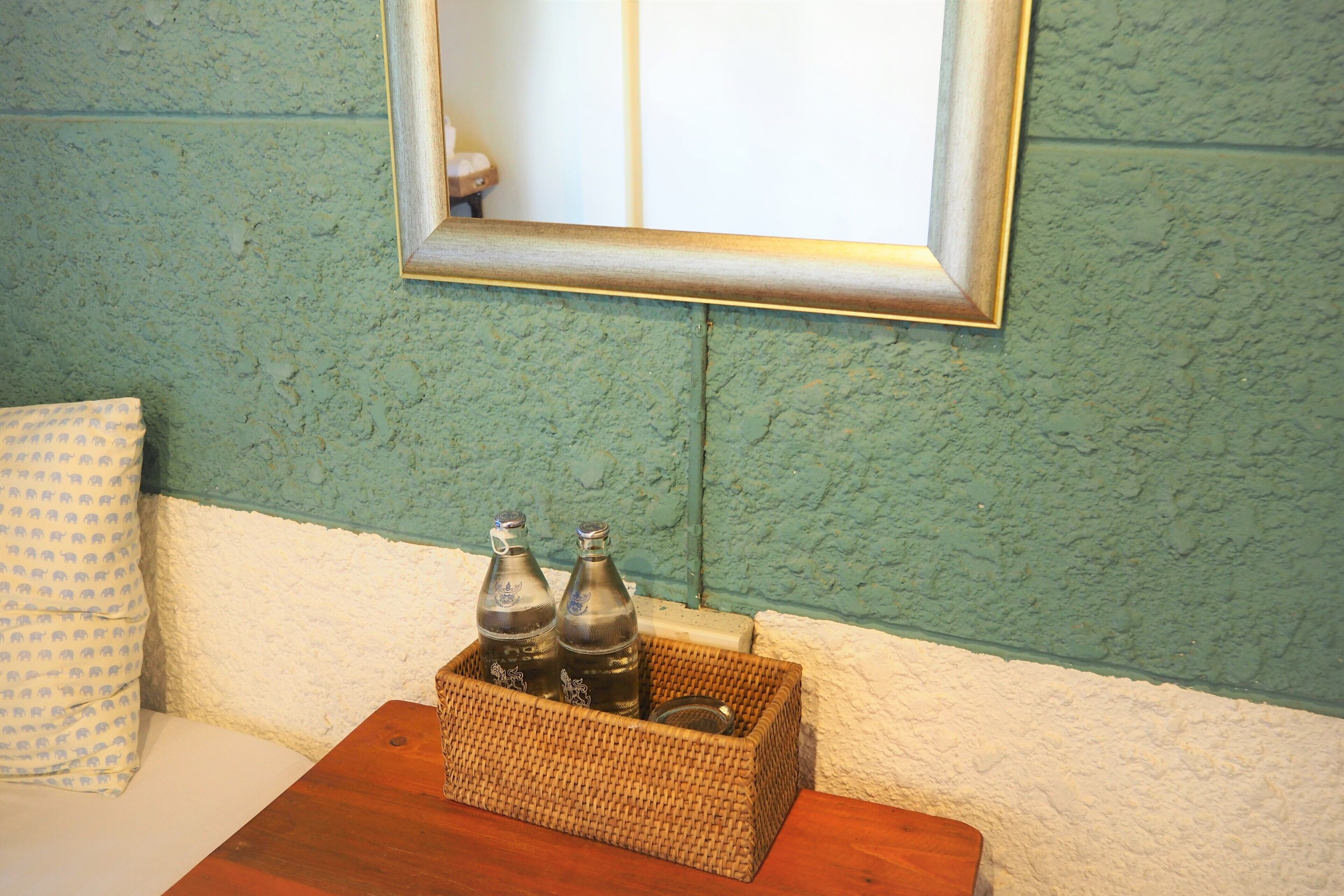 A room with a bed, a small table with a wicker basket containing two bottles, and a mirror on the wall.