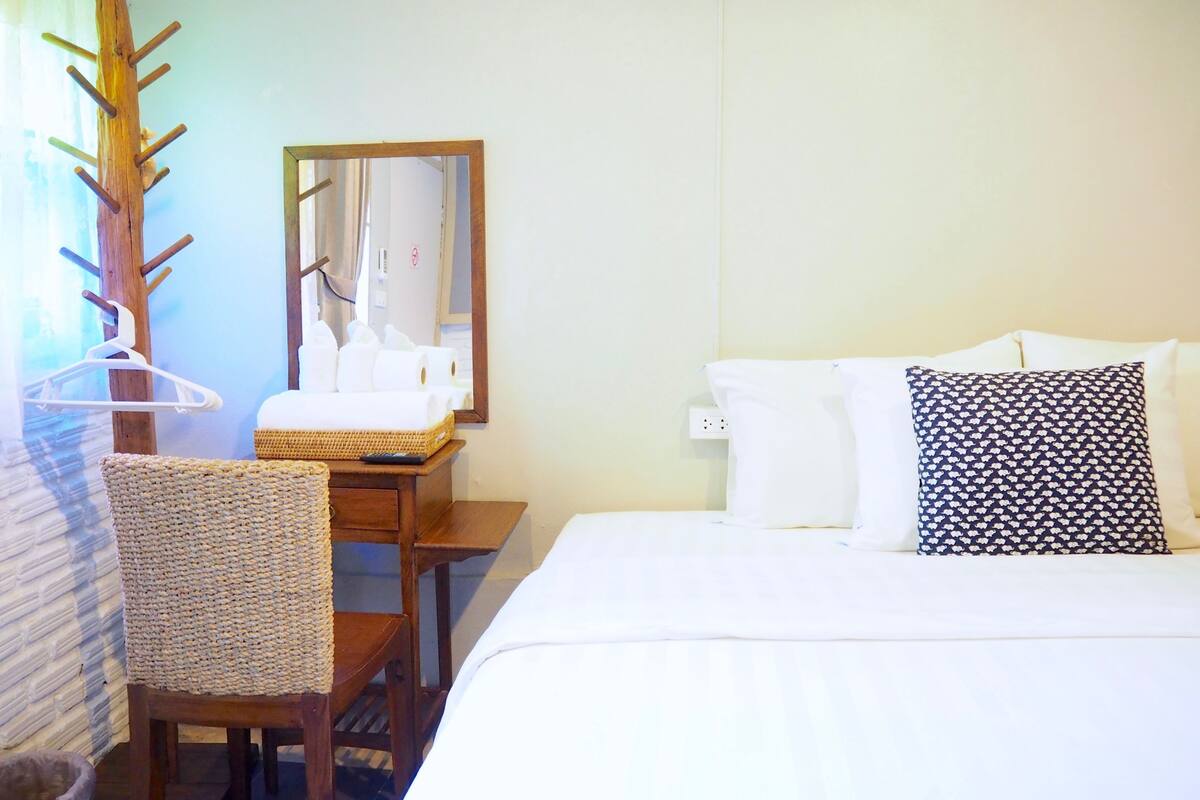 A hotel room with a bed, a wooden chair, a small table, a mirror, and a hanging coat rack.