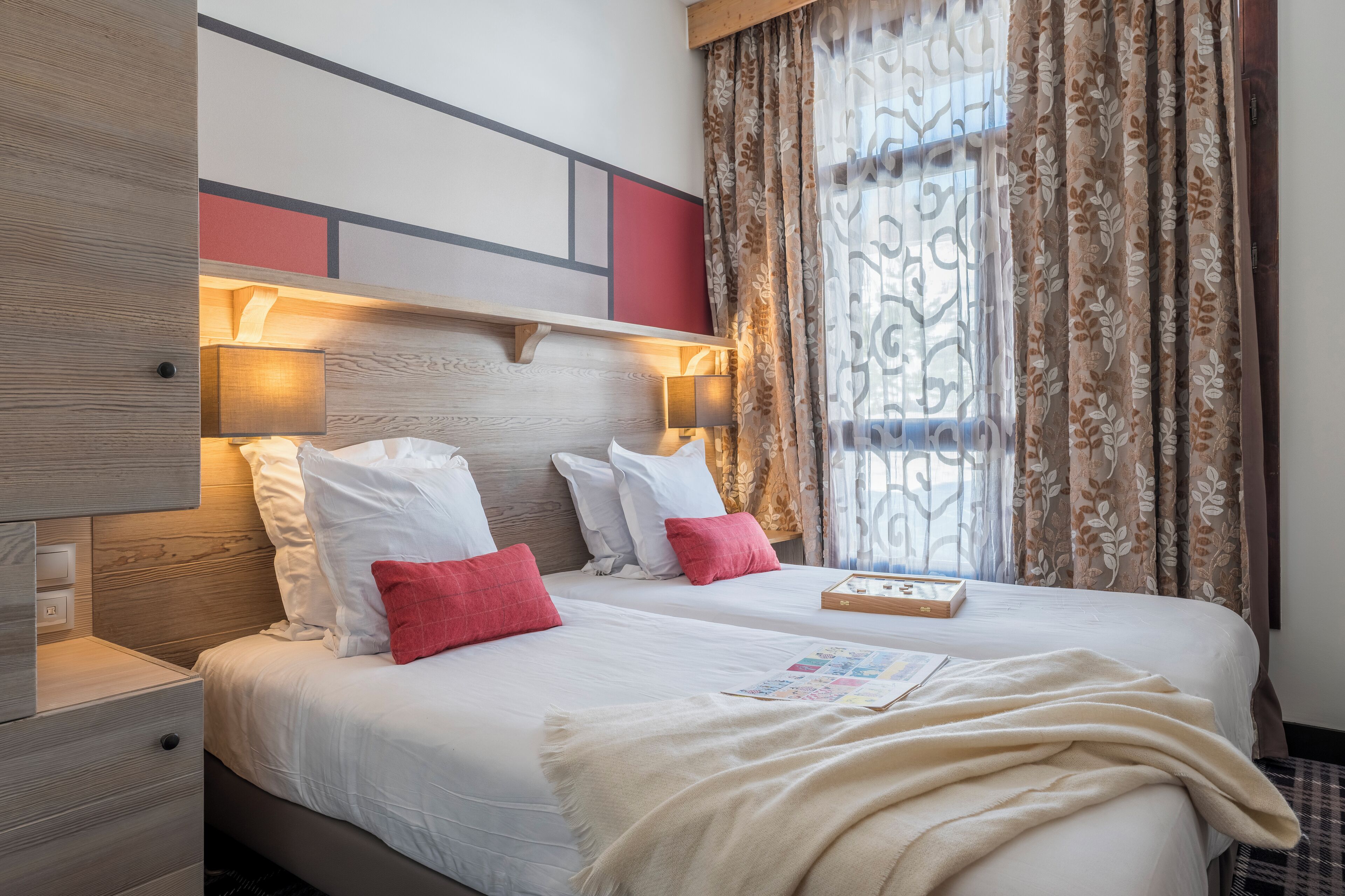 Un lit double avec des oreillers blancs et rouges, un plaid beige et une table de chevet avec une lampe.