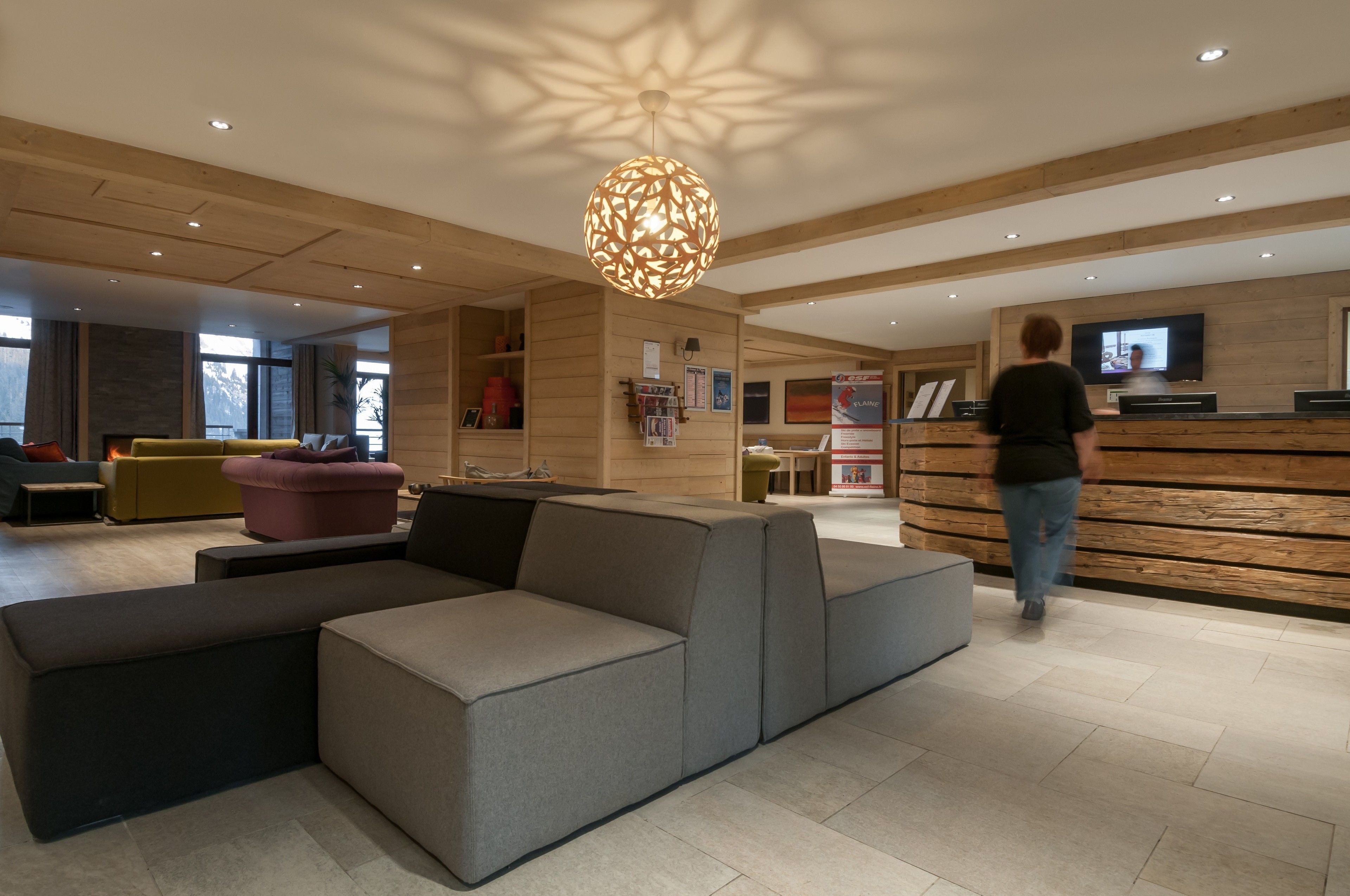 Un hall d’hôtel moderne avec un grand canapé d’angle, un comptoir d’accueil en bois et un grand luminaire suspendu.