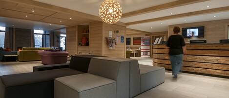 Un hall d’hôtel moderne avec un grand canapé d’angle, un comptoir d’accueil en bois et un grand luminaire suspendu.