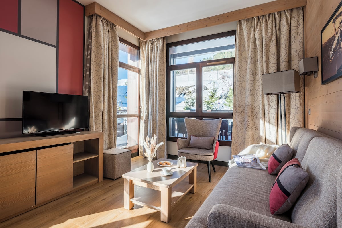 A modern living room with a flat-screen TV, wooden furniture, a sofa with cushions, a coffee table, and large windows with curtains.