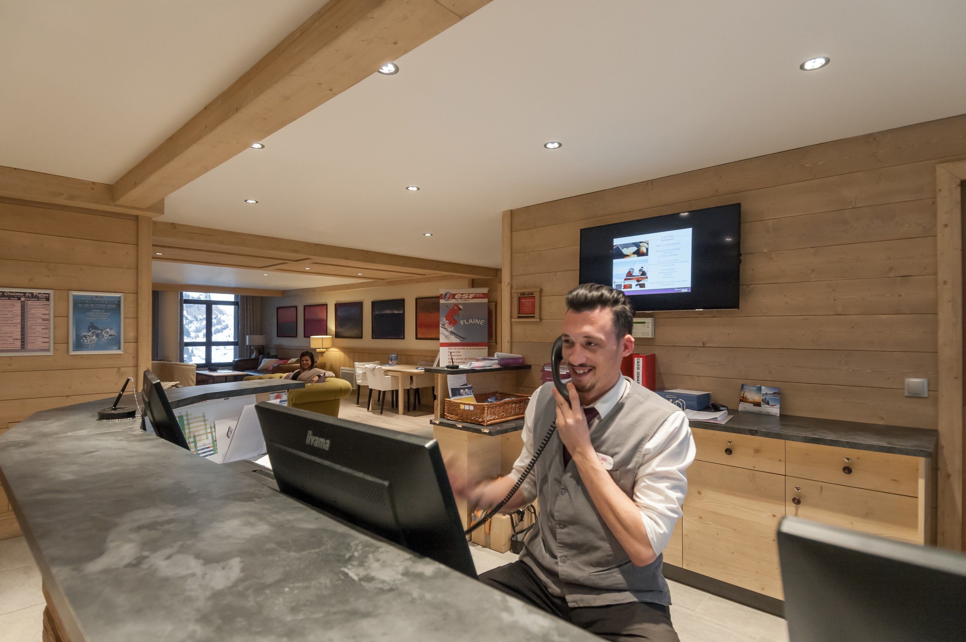 Un espace d’accueil d’hôtel avec un intérieur en bois, un téléviseur à écran plat fixé au mur et un bureau équipé d’un ordinateur et d’un téléphone.