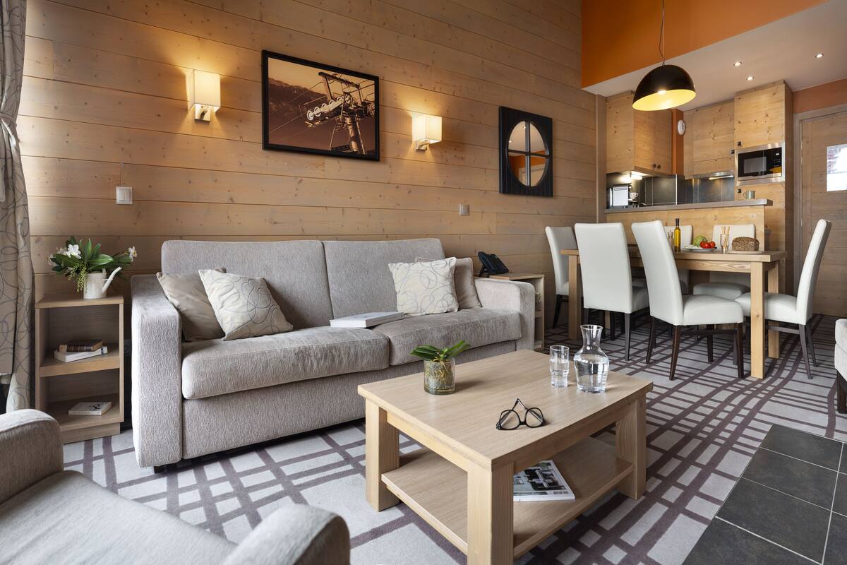 A modern living room with a sofa, coffee table, and chairs. There is a dining area with a table and chairs. The room has wooden walls and a kitchen area with a microwave and oven.
