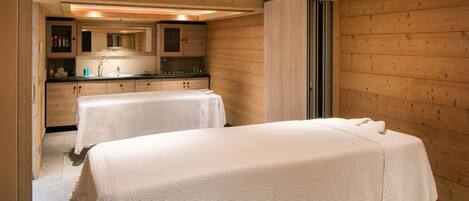 A room with two beds covered in white linens, wooden walls, a wooden ceiling, and a kitchen area with cabinets and a countertop.
