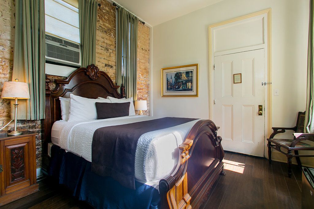 Een hotelkamer met een houten bed, nachtkastje, stoel, deur en een ingelijst schilderij aan de muur.