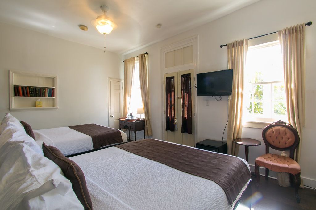 Una habitación de hotel con dos camas, televisión, estantería, ventana con cortinas y una silla.