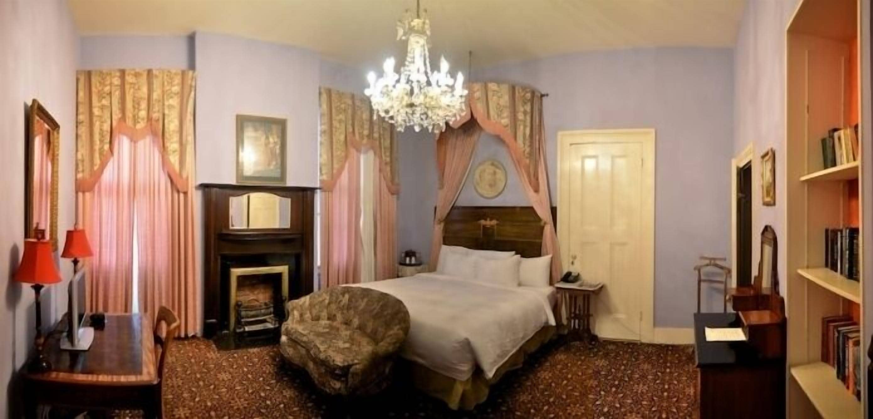 A bedroom with a bed, a fireplace, a chandelier, curtains, a bookshelf, and a small table.
