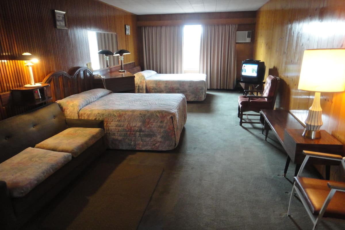 A hotel room with a sofa, two single beds, a TV, a desk with a lamp, and a chair.