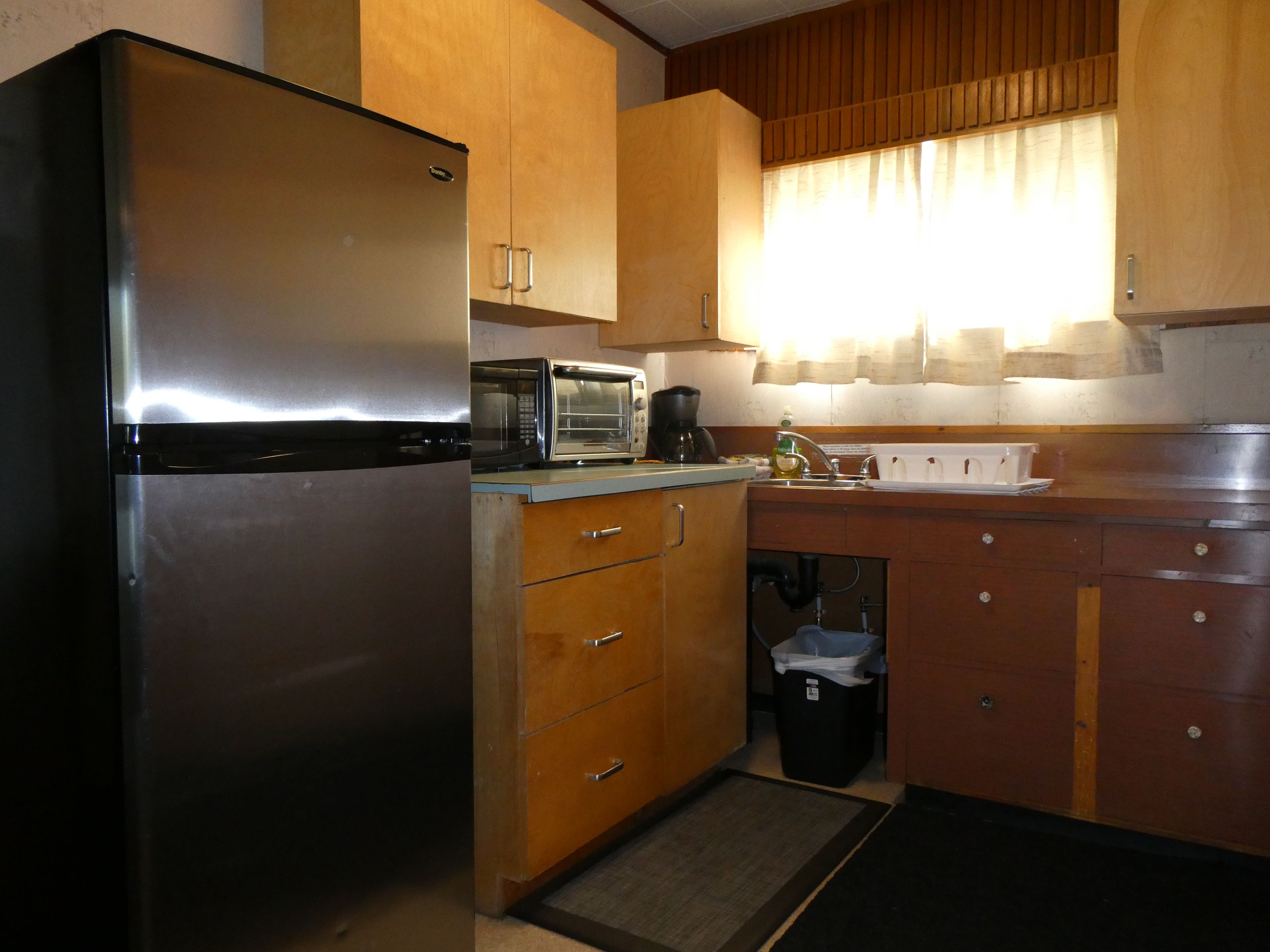 A compact kitchen with a stainless steel refrigerator, a microwave, a coffee maker, wooden cabinets, and a small trash bin.
