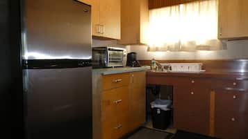 A compact kitchen with a stainless steel refrigerator, a microwave, a coffee maker, wooden cabinets, and a small trash bin.