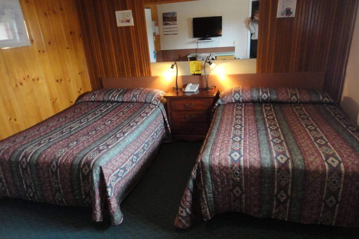 A hotel room with two single beds, a wooden headboard, a bedside table with a lamp, a telephone, and a flat-screen TV mounted on the wall.