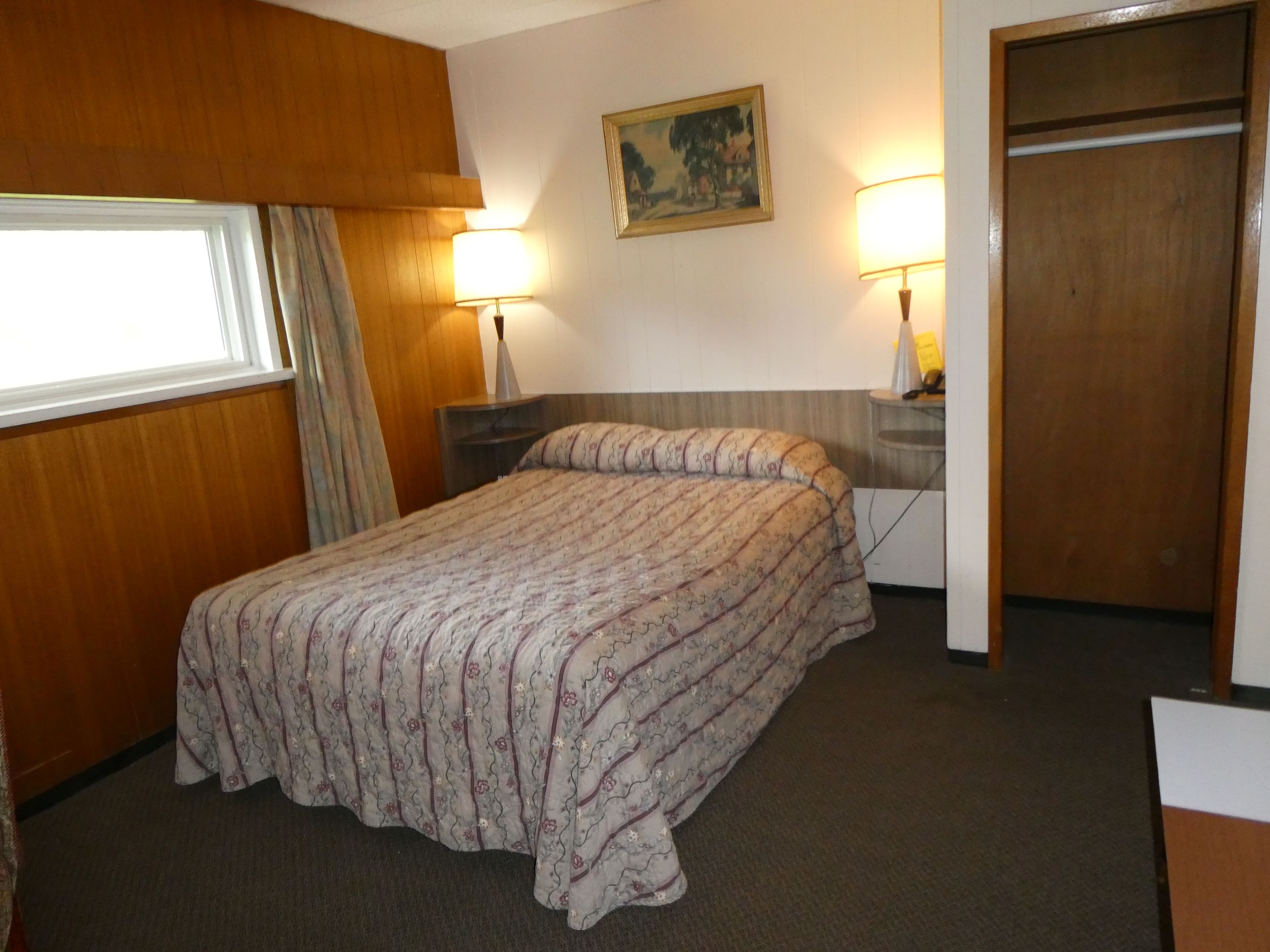 A hotel room with a bed, two bedside lamps, a painting on the wall, and a wooden wardrobe.