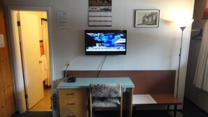 Una habitación con escritorio, una silla, televisor montado en la pared, lámpara y puerta que da a otra habitación.