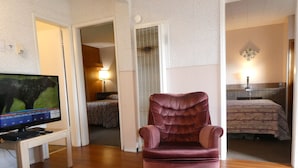A room with a wooden floor, a television on a stand, a plush armchair, a bed with a patterned bedspread, and a wall-mounted lamp. - Airport Inn Motel & RV Park (Quesnel)