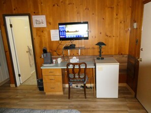 A small, compact room with a wooden paneled wall, a desk with a computer, a chair, a small refrigerator, and a coffee maker.