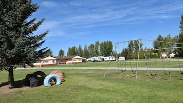 A grassy field with a swing set, a tree, and RVs in the background.