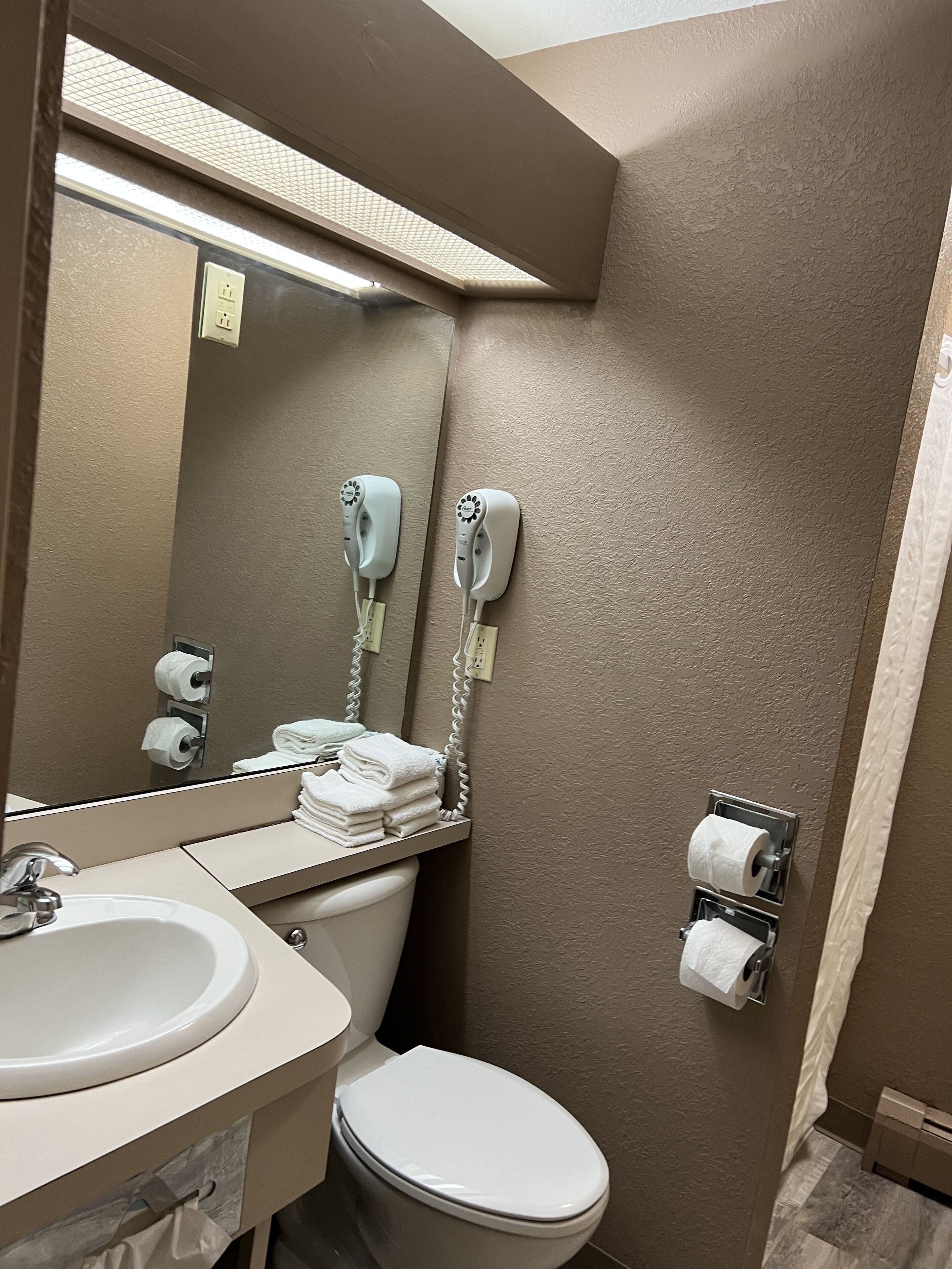 A bathroom with a sink, mirror, toilet, towel dispenser, and hair dryer.