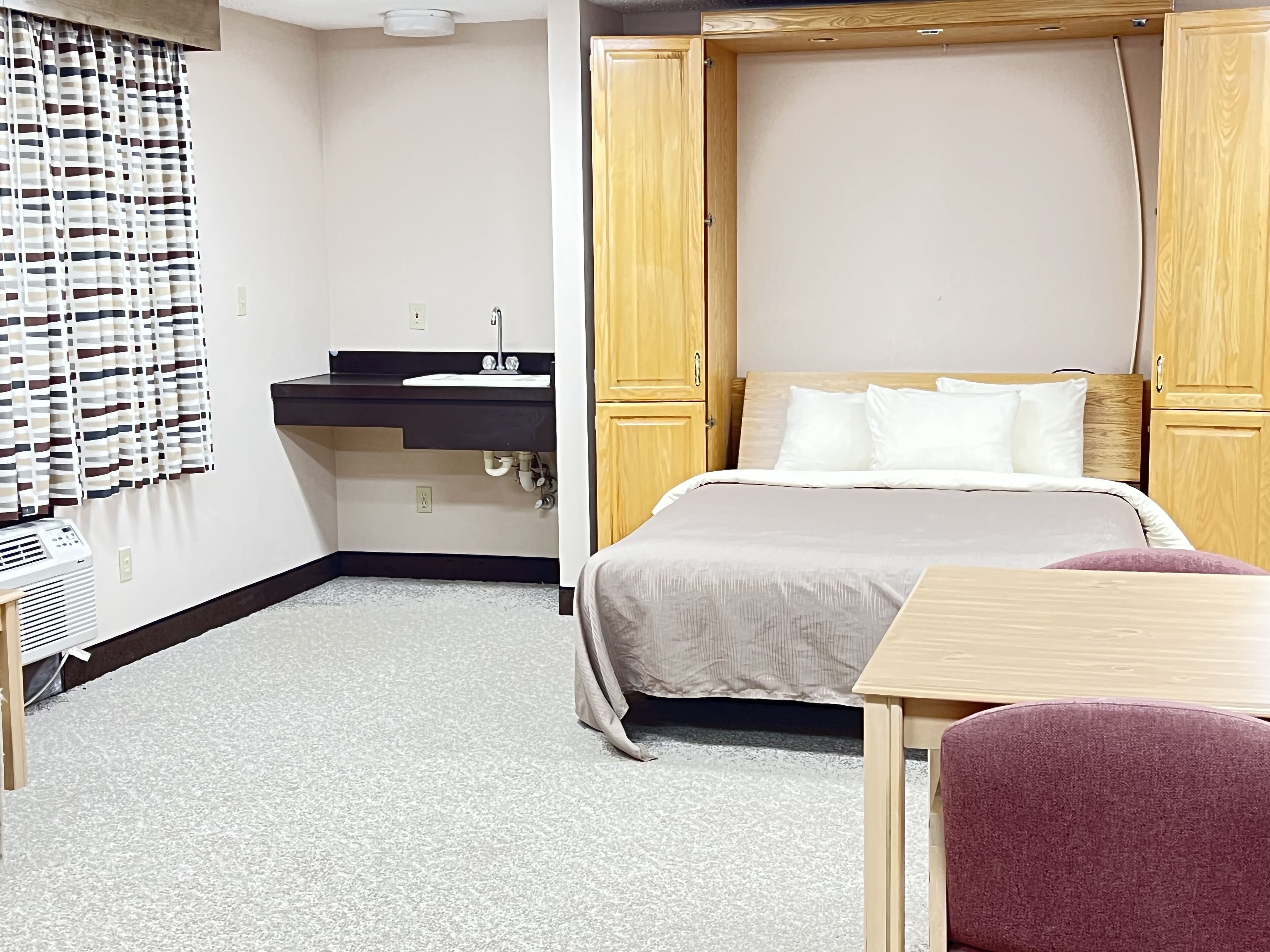 A hotel room with a bed, a bedside table, a wooden wardrobe, a bathroom with a sink, and a patterned curtain.