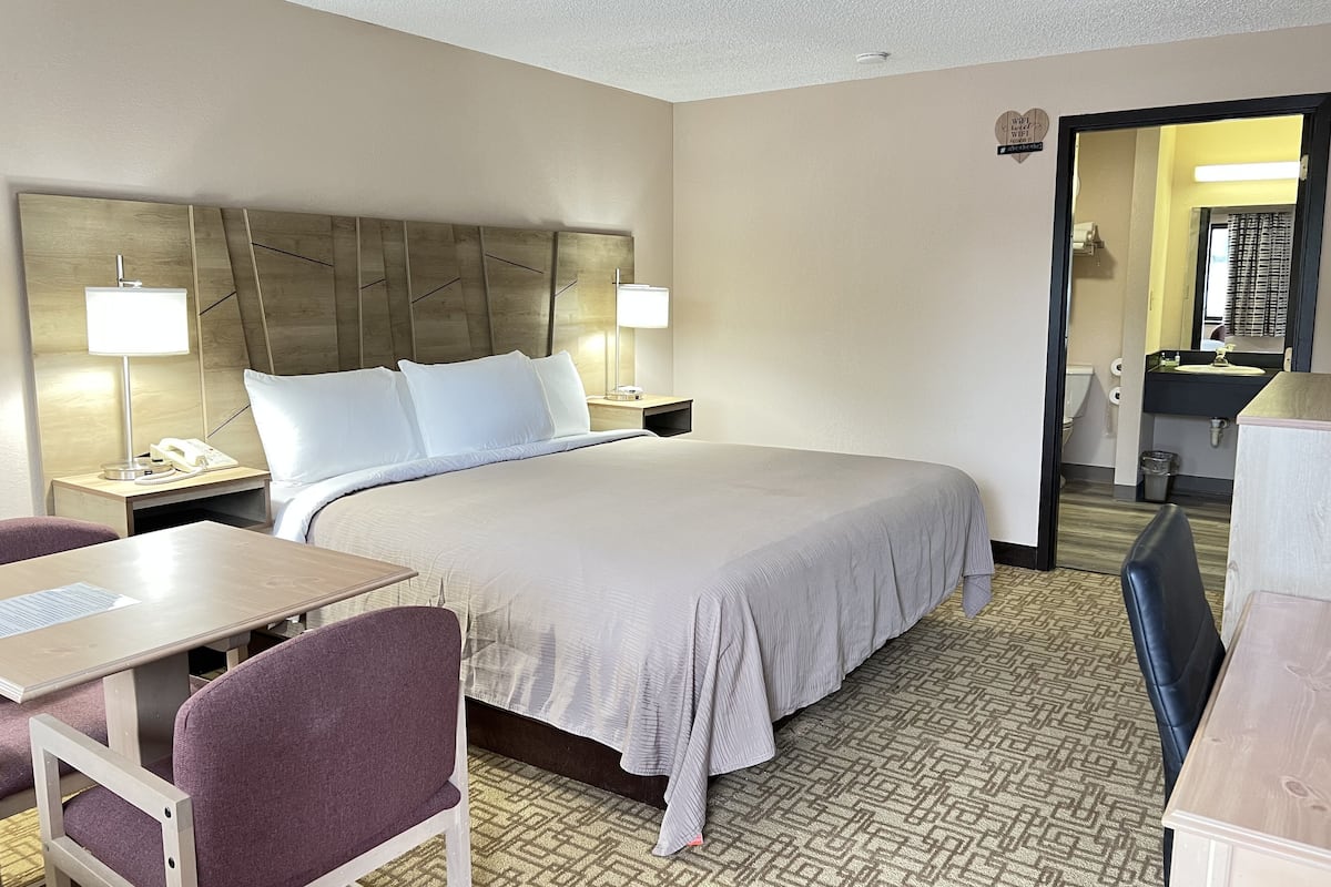 A hotel room with a large bed, bedside tables with lamps, a desk, a chair, and a bathroom visible through an open door.
