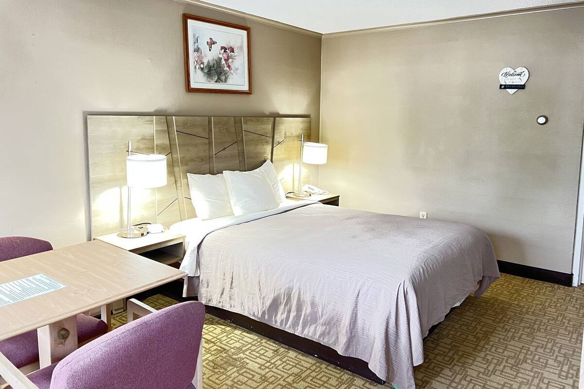 A hotel room with a bed, bedside lamps, a desk, a chair, a framed picture on the wall, and a welcome sign.