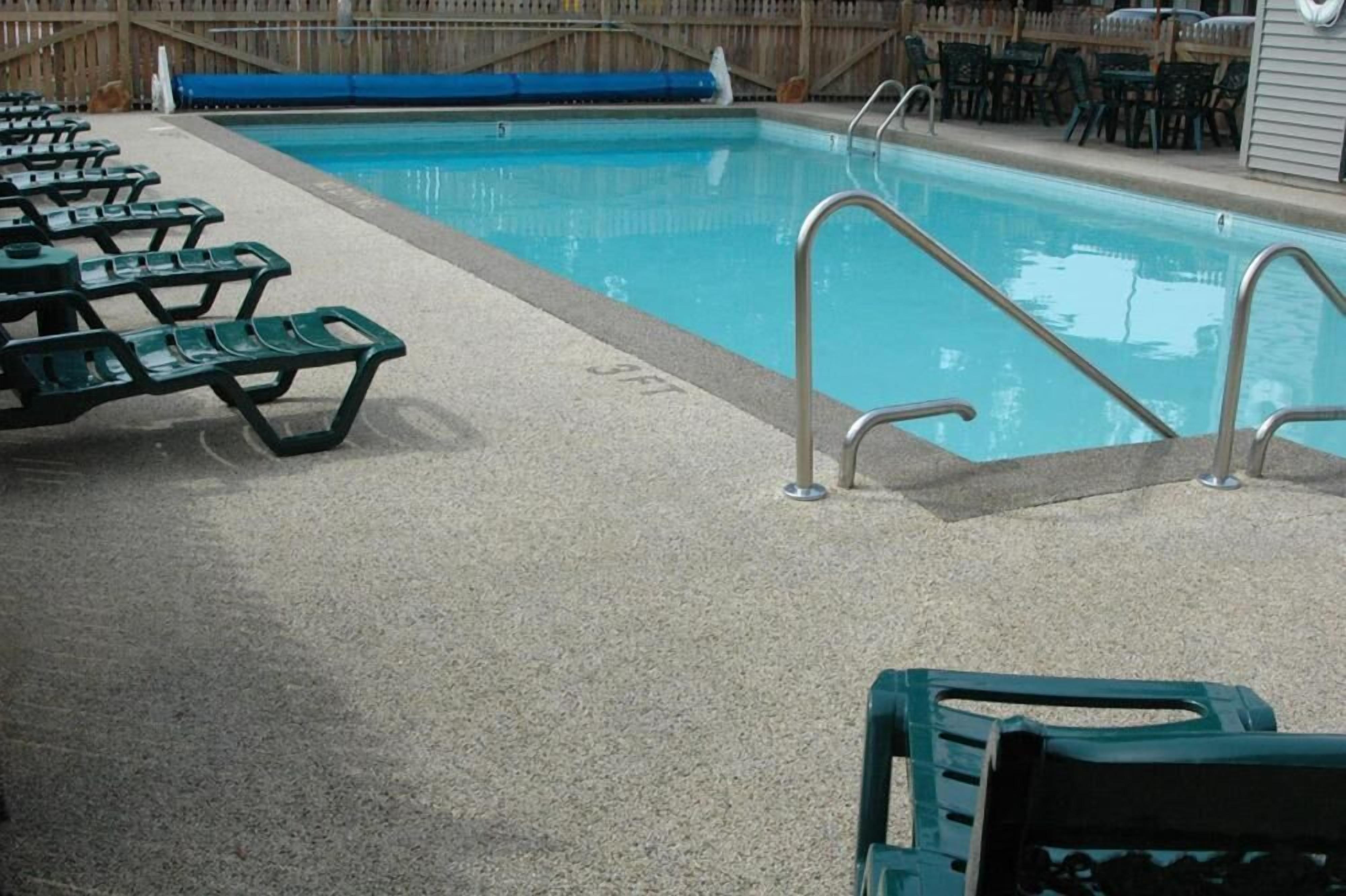A swimming pool with a concrete deck, metal pool ladder, and green lounge chairs.