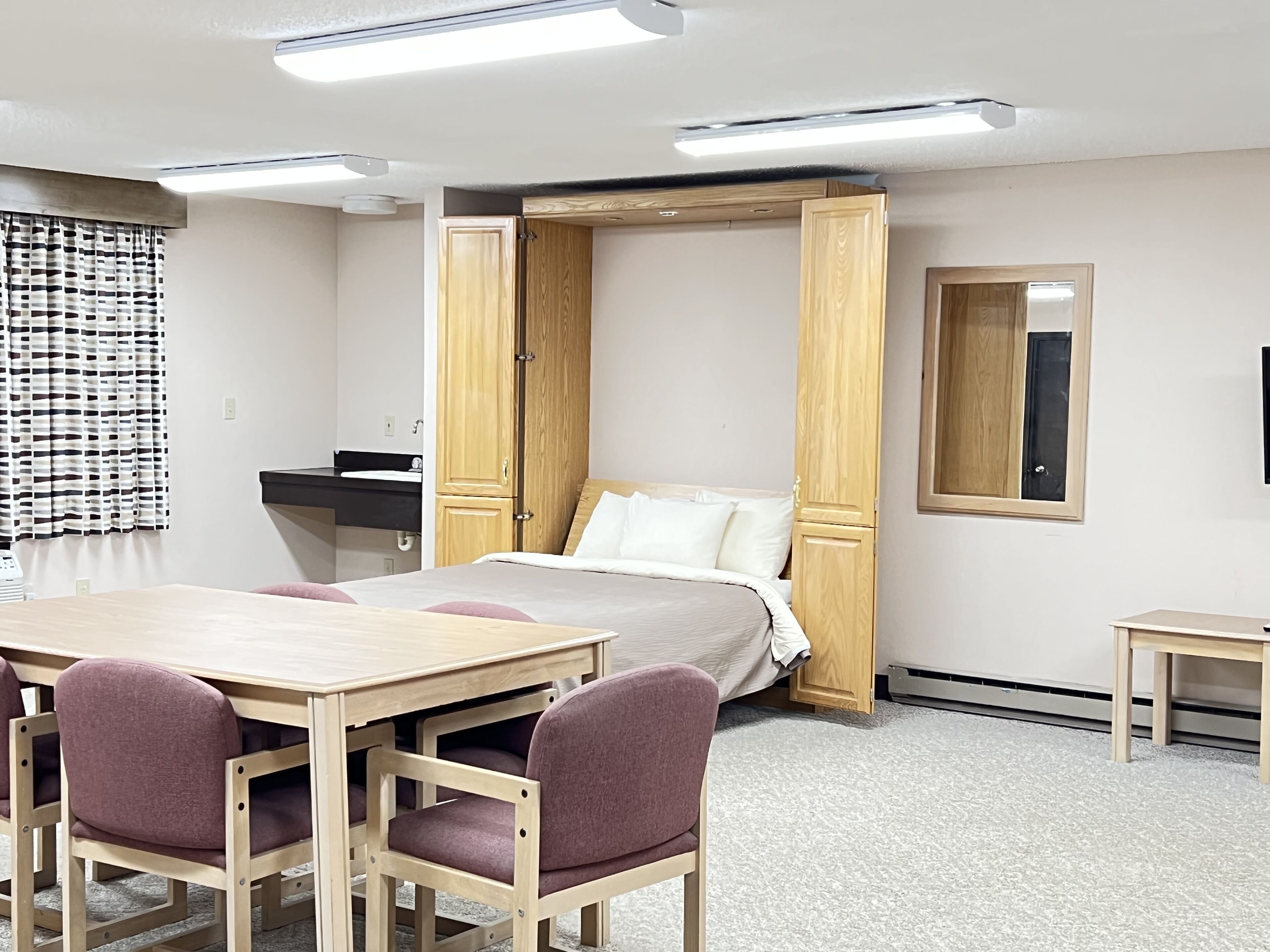 A room with a bed, a desk, and chairs. There is a mirror on the wall and a window with curtains.