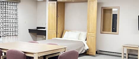 A room with a bed, a desk, and chairs. There is a mirror on the wall and a window with curtains.