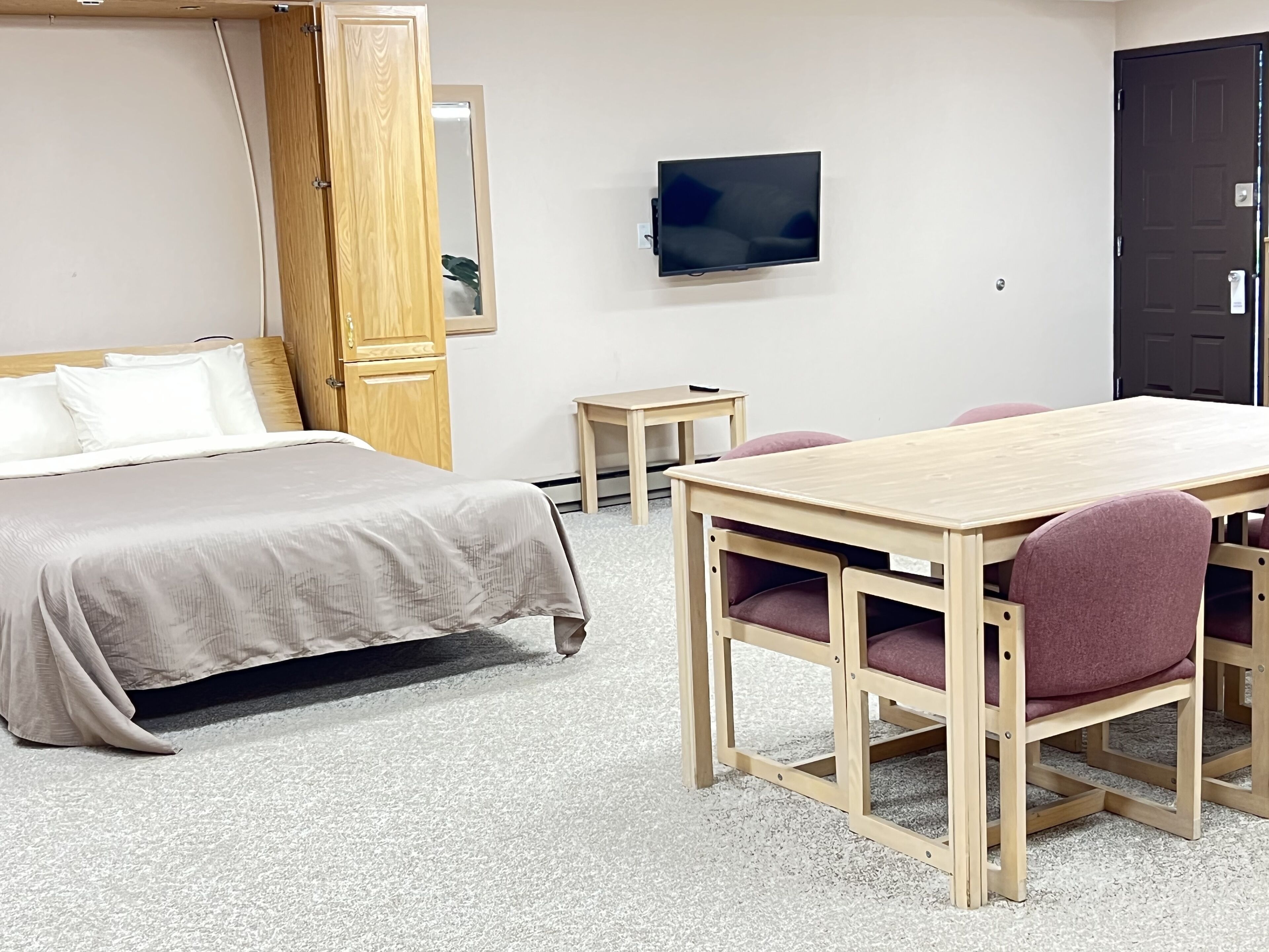 Une chambre d’hôtel avec un lit, une table en bois avec des chaises, un téléviseur fixé au mur et une porte.