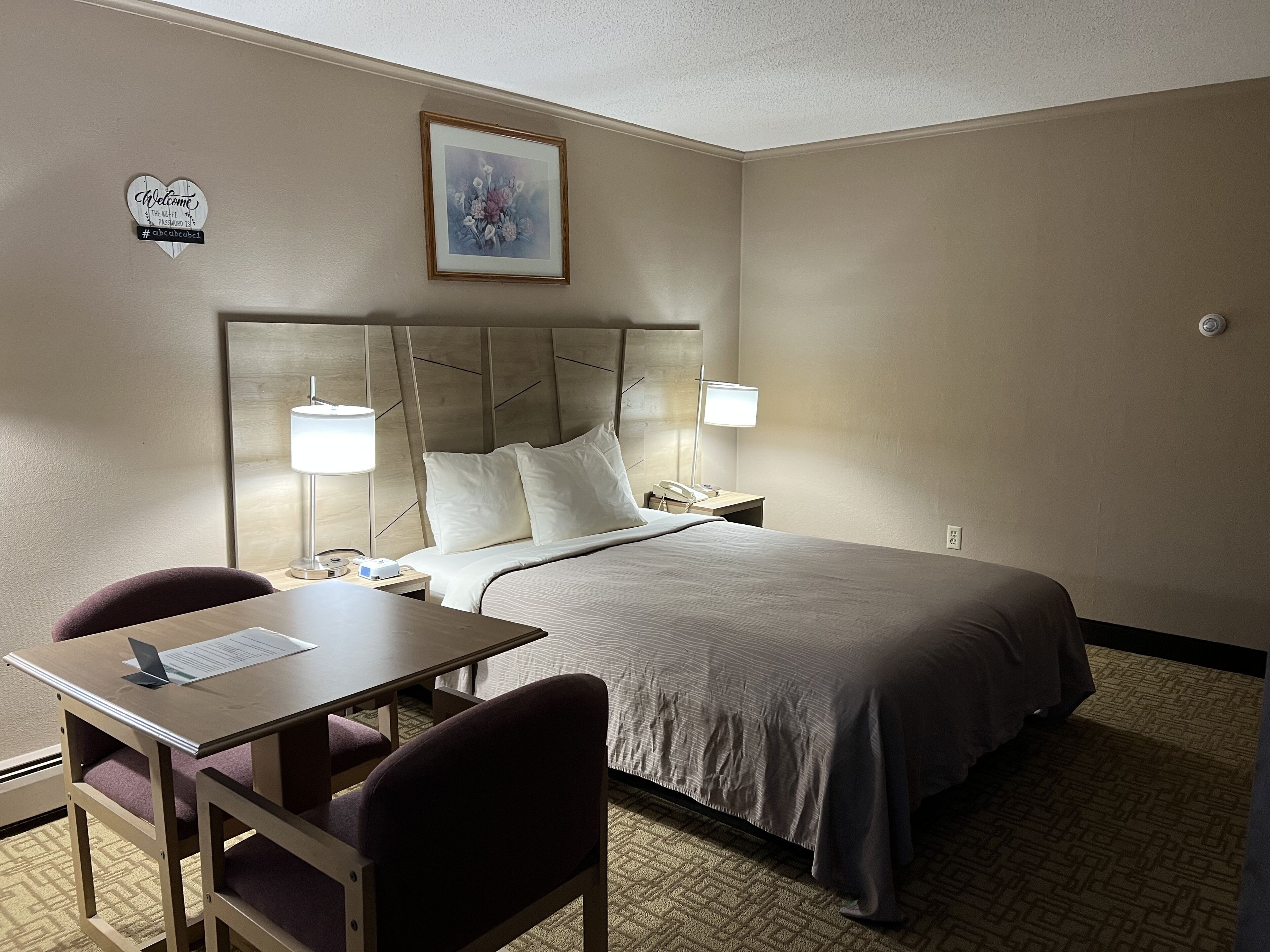 Quarto de hotel com cama, luminárias de cabeceira, escrivaninha e uma cadeira, e um letreiro de boas-vindas emoldurado.