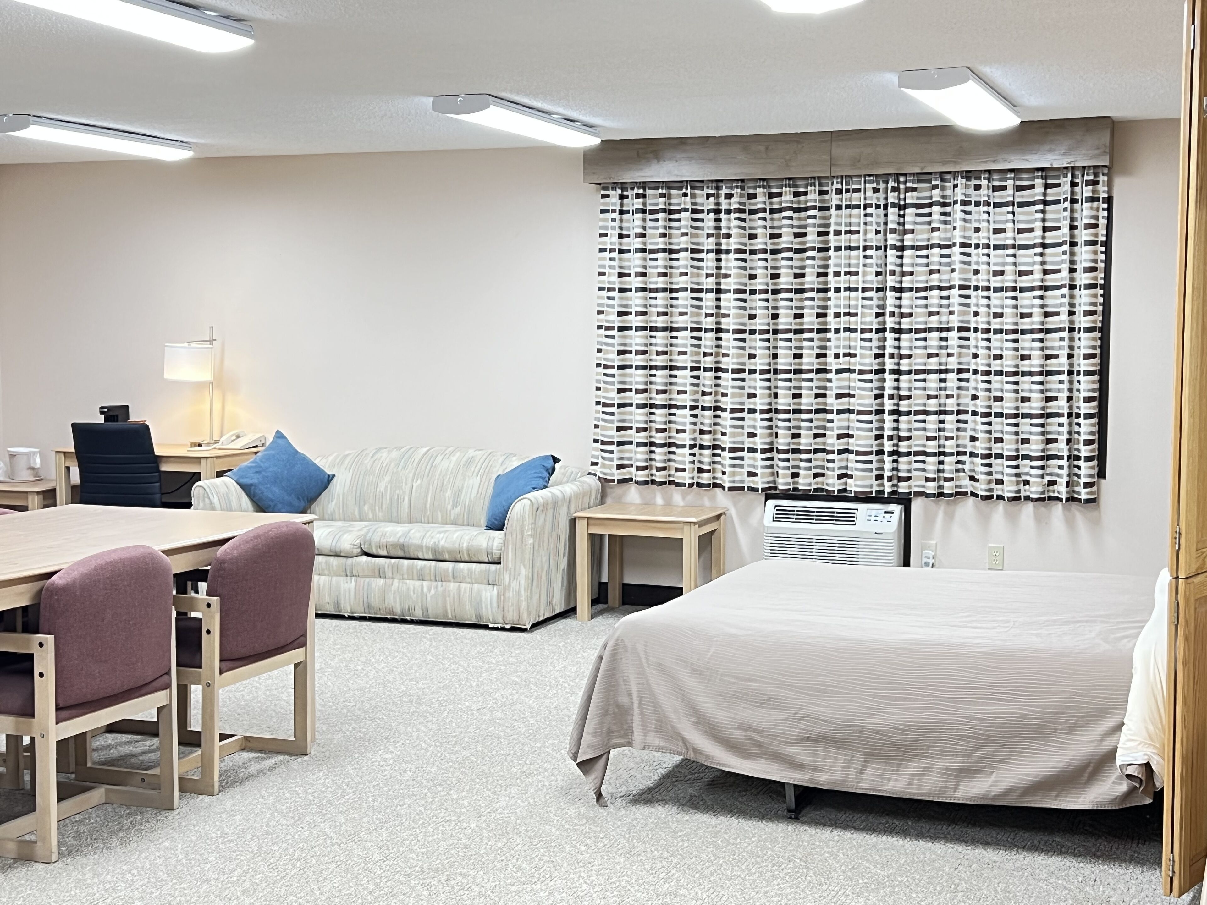 A hotel room with a bed, a desk, a chair, a sofa, a lamp, and a window with patterned curtains.