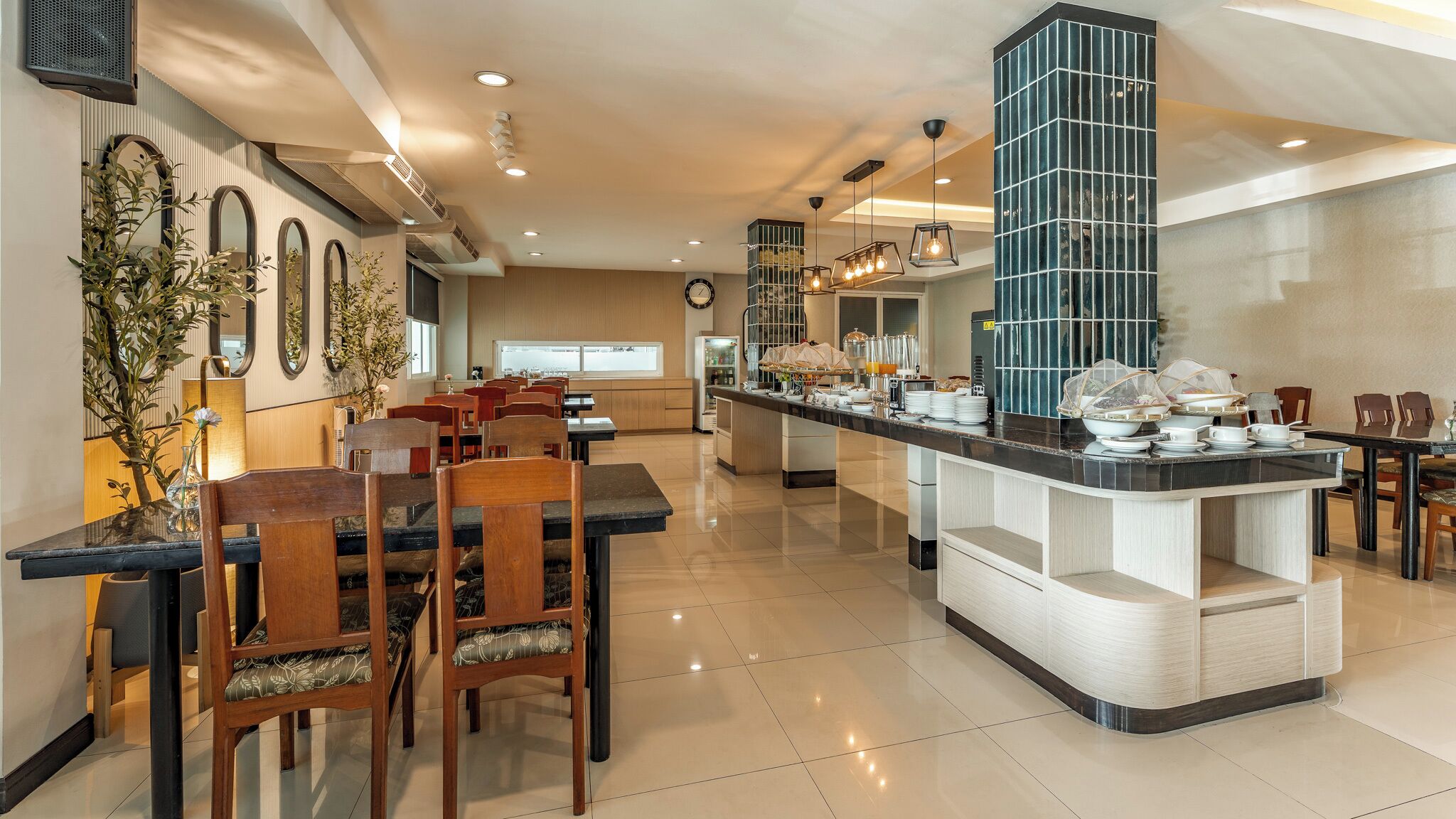 A modern hotel dining area with a central island, wooden chairs, and a long counter with breakfast items.