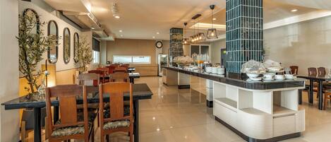 A modern hotel dining area with a central island, wooden chairs, and a long counter with breakfast items.