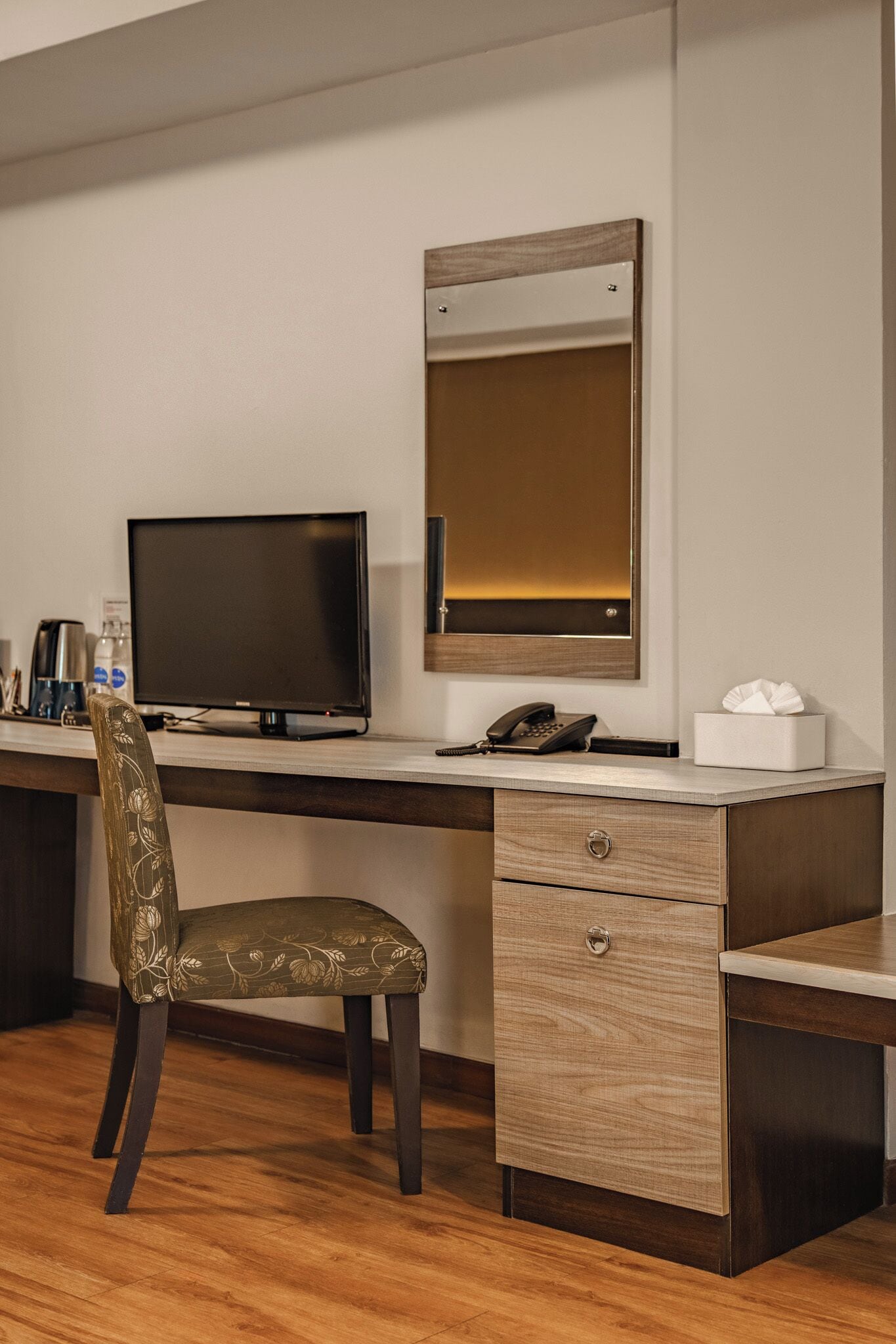 A hotel room with a wooden desk, a television, a telephone, a chair, and a bedside table with a tissue box.