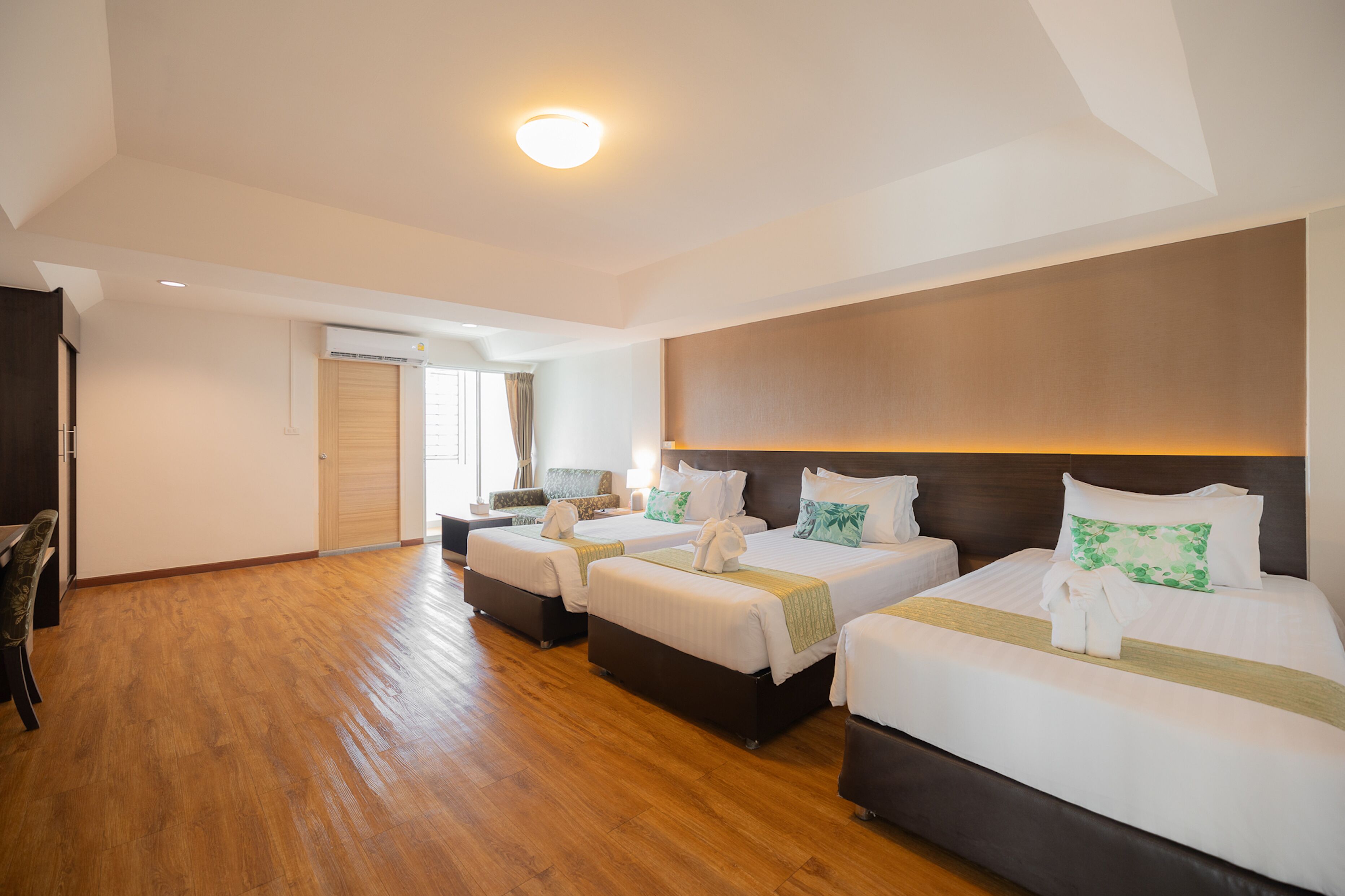 A hotel room with three beds, wooden flooring, a large window, and a modern ceiling light.