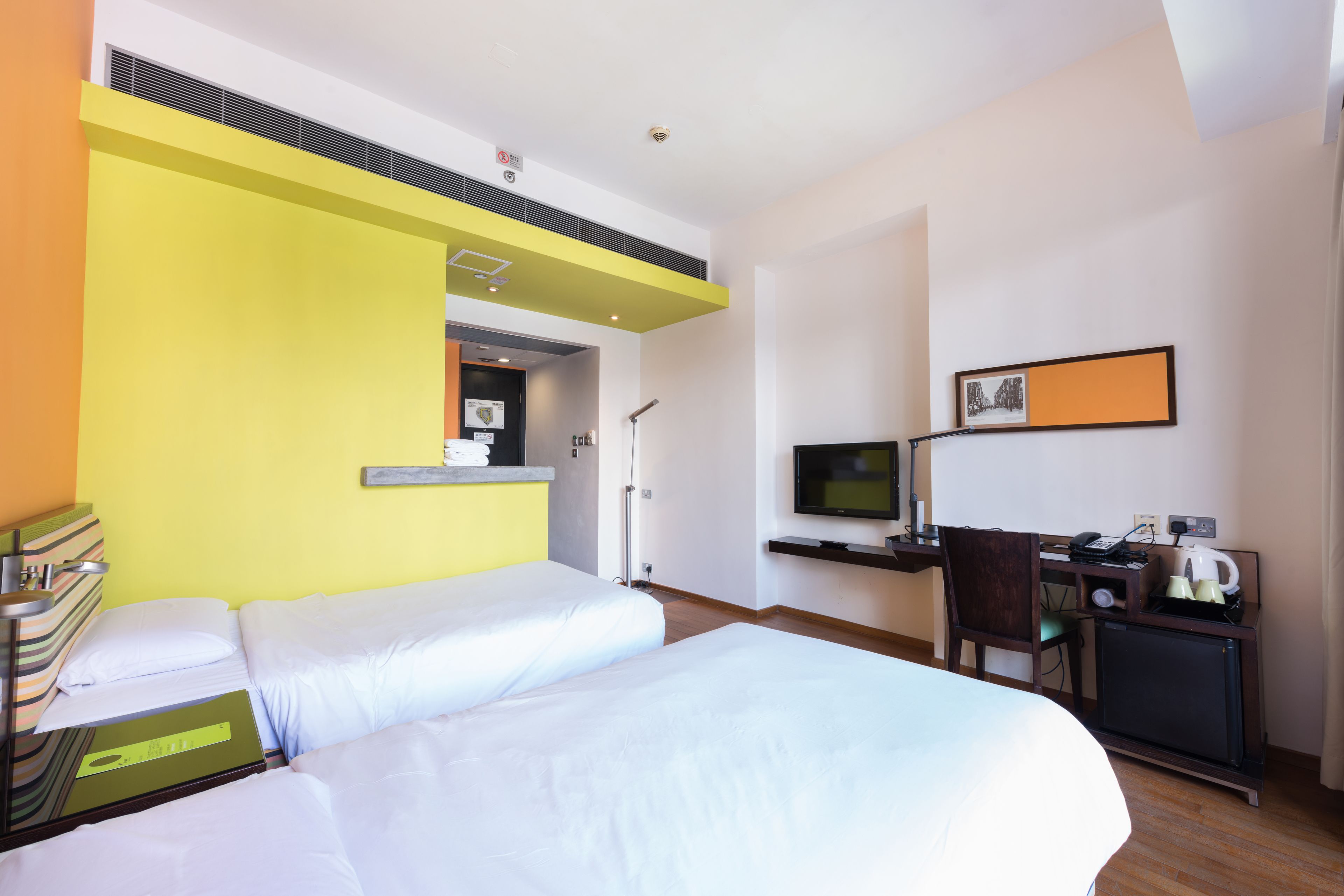 A hotel room with two beds, a desk with a computer, a TV, and a small kitchenette.