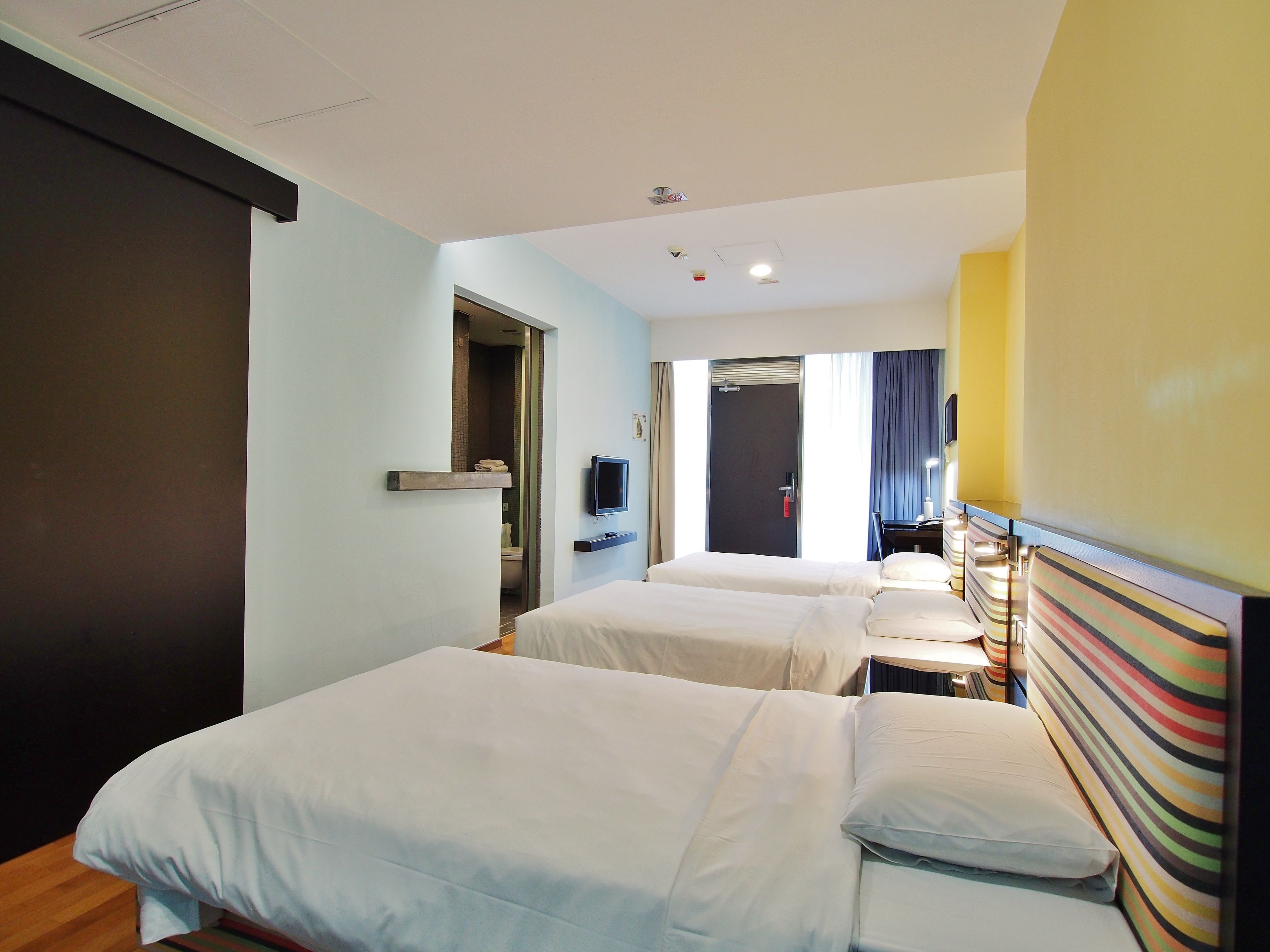 Quarto de hotel com duas camas, uma televisão, banheiro visível por uma porta aberta e uma janela com cortinas.