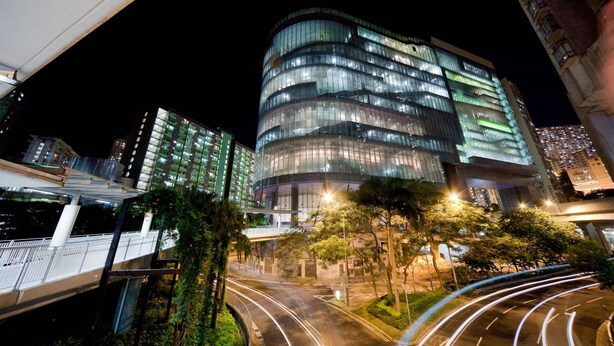 Un edificio moderno con fachada curva y varios pisos, rodeado de vegetación y un puente peatonal.