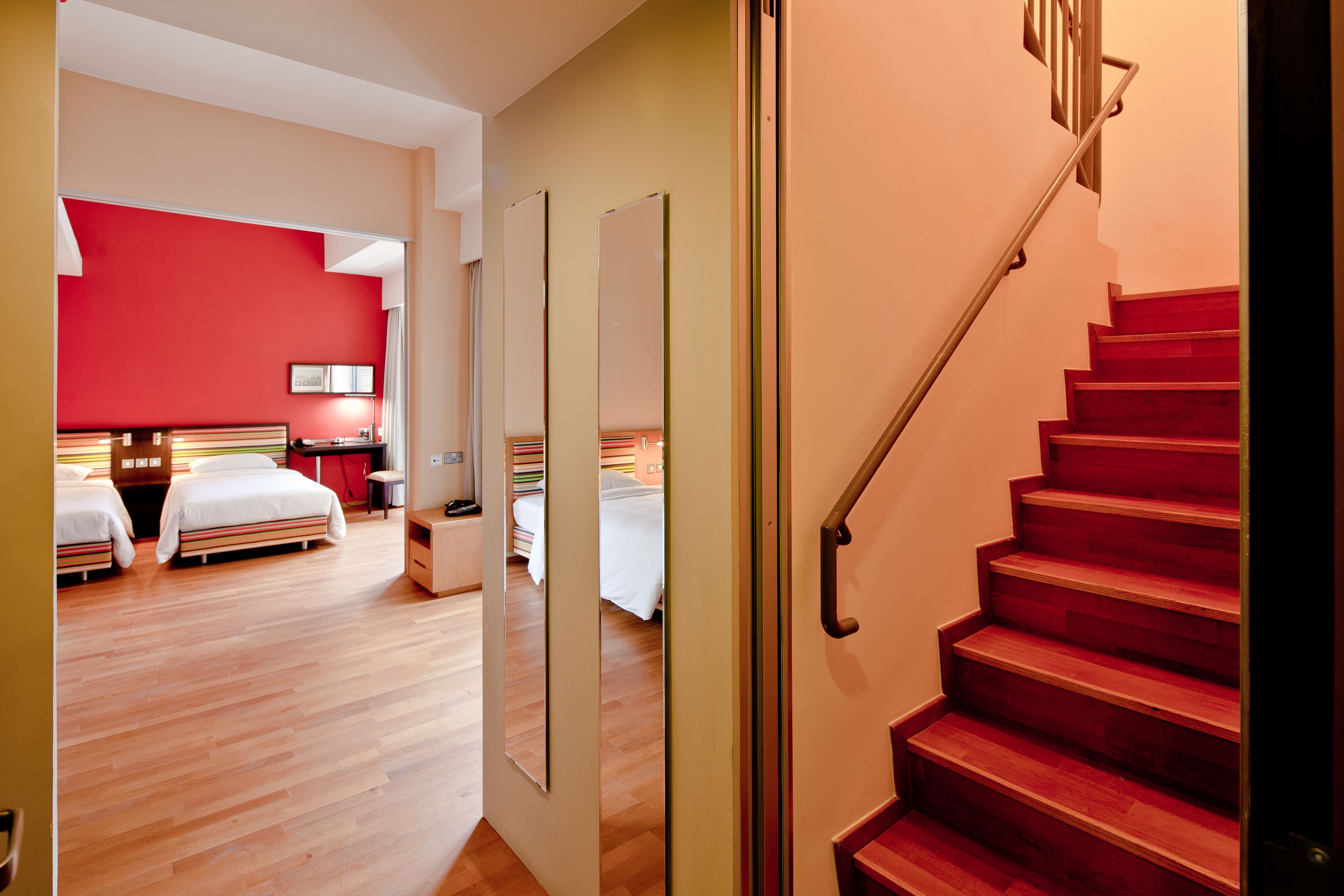 A hotel room with a wooden floor, a red wall, and a staircase with a metal handrail.