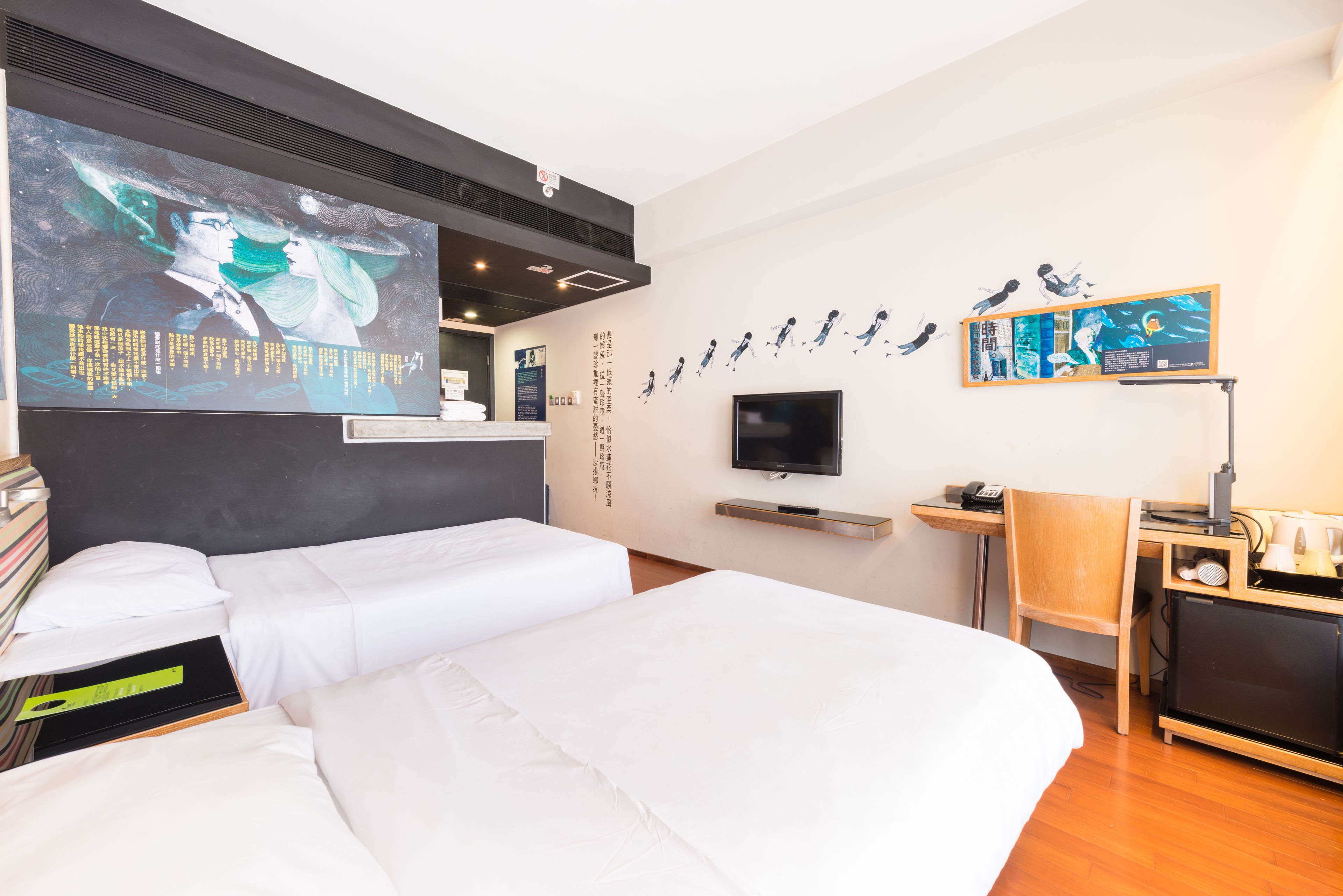 Una camera d'albergo con due letti, una scrivania con un computer, una televisione e opere d'arte alle pareti.