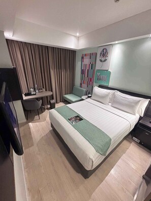 Une chambre d’hôtel avec un lit, un bureau, une chaise, une télévision et un mur orné d’une œuvre d’art colorée.