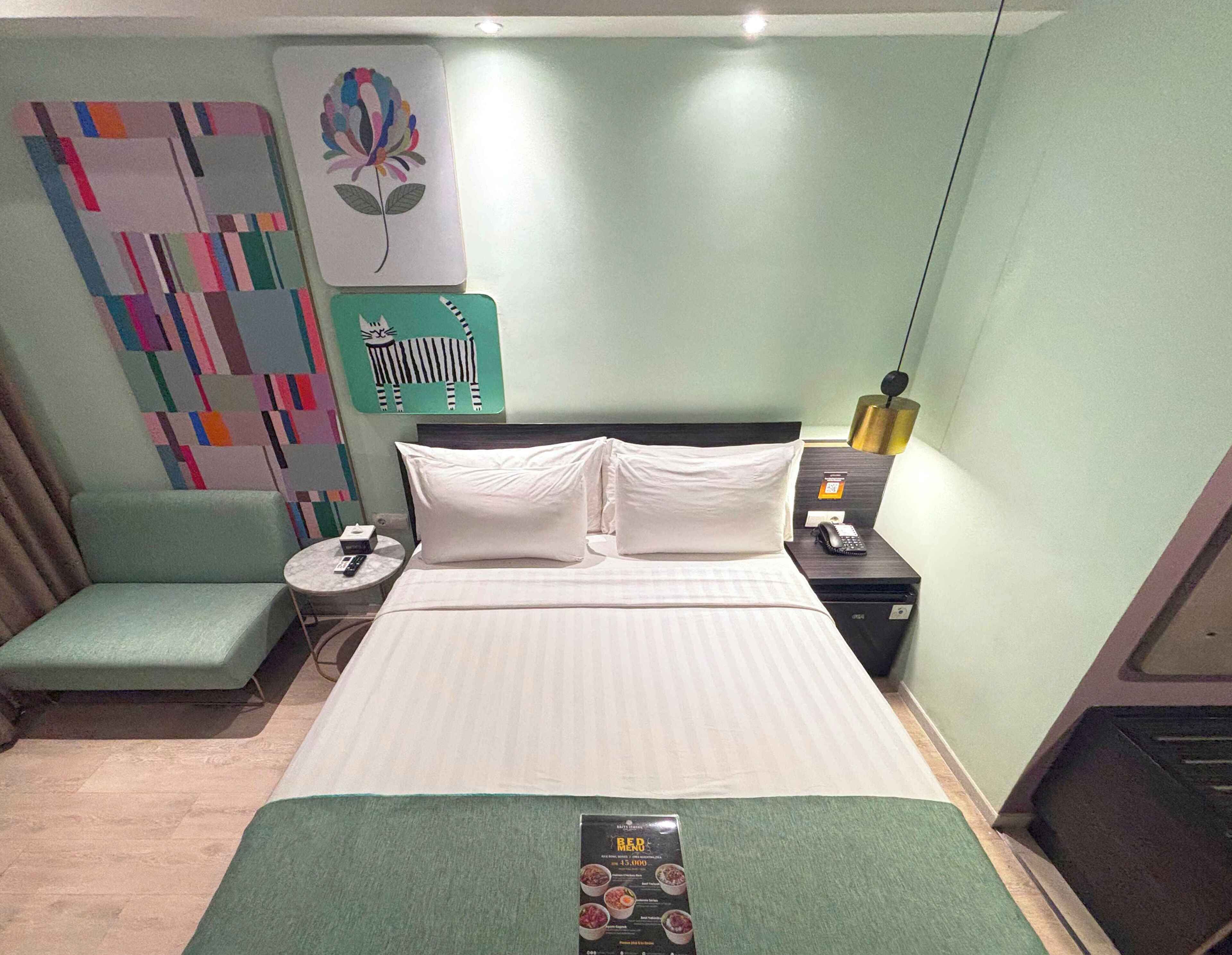 A hotel room with a bed, a bedside table, a chair, a wall with a colorful abstract painting, and a small table with a menu.