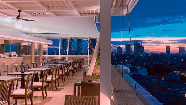 Restaurante no terraço com mesas e cadeiras, um ventilador de teto e vista para a cidade ao entardecer.