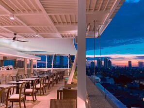 Un restaurant situé sur un toit, avec des tables et des chaises, un ventilateur de plafond et une vue sur la ville au crépuscule.