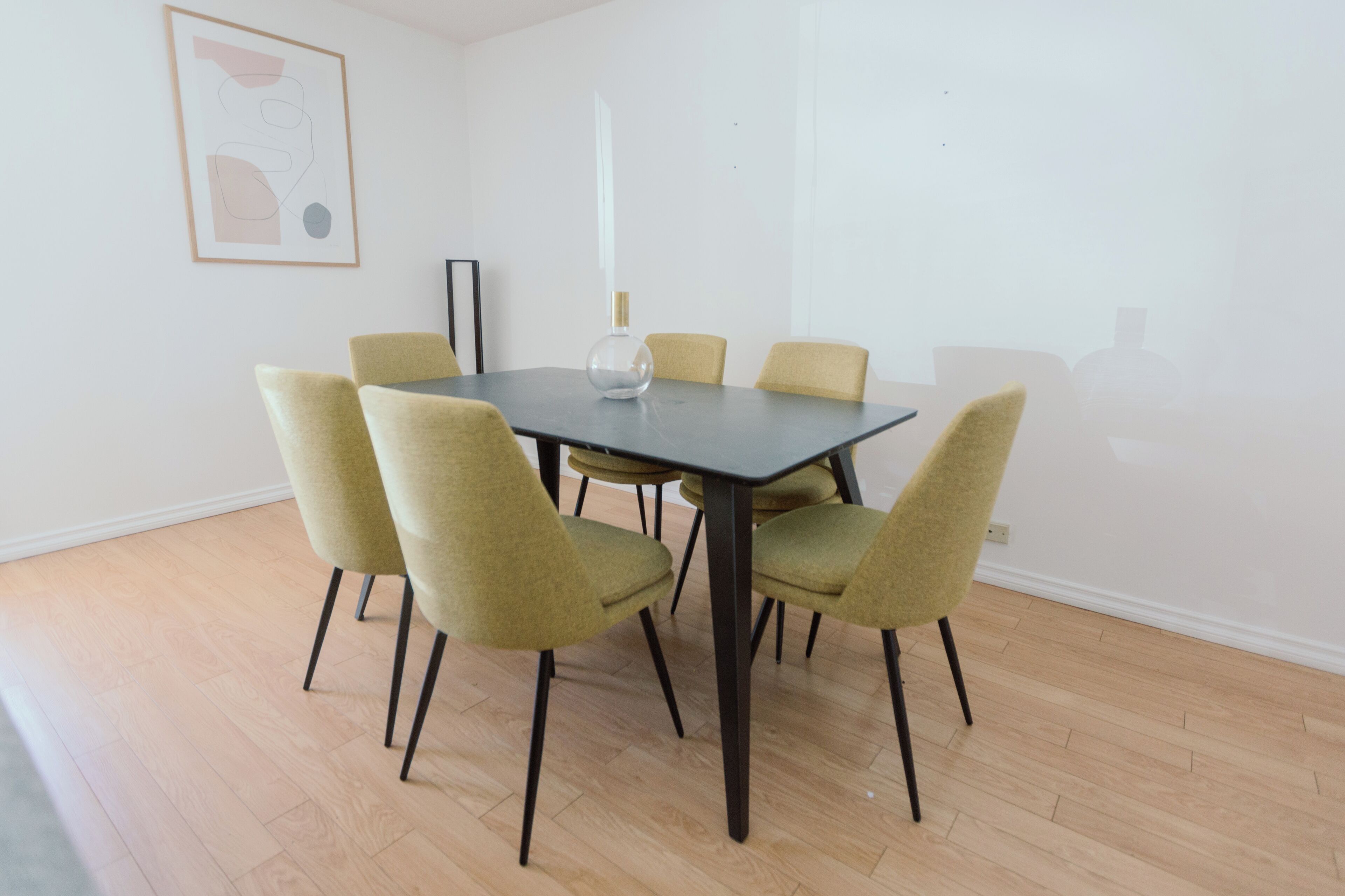 A dining area with a rectangular table surrounded by six chairs, a vase on the table, and a framed abstract artwork on the wall.