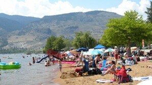 A beach with people sunbathing and swimming, a playground, and a mountain backdrop. - Waterfront Inn (Penticton)