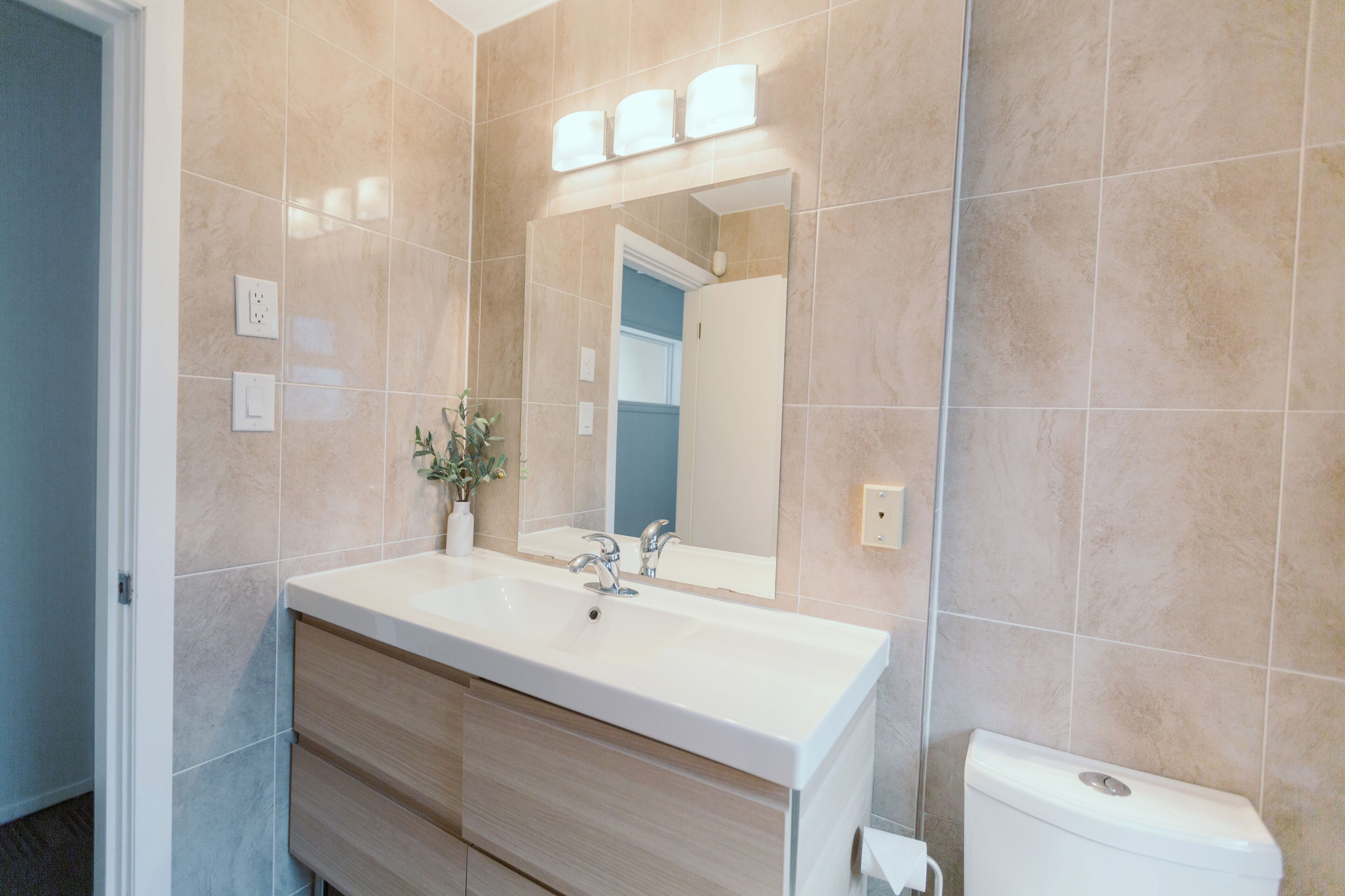 A bathroom with a beige tiled wall, a white sink with a mirror above it, and a toilet to the right.